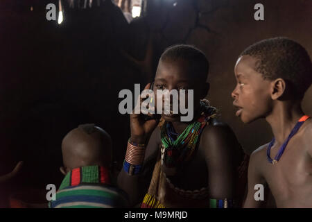 Giovane donna con bambini, telefono cellulare le chiamate nella capanna di fango, Hamer tribe, Turmi, Regione del Sud unite, Etiopia Foto Stock