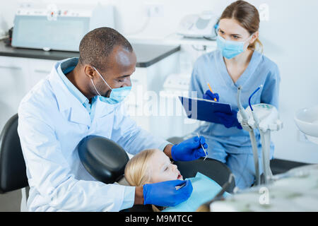 Note di vitale importanza. Affascinante femmina giovane infermiere rendendo note di dentisti osservazioni mentre stava svolgendo un accurato checkup di un po' di ragazze della cavità orale Foto Stock