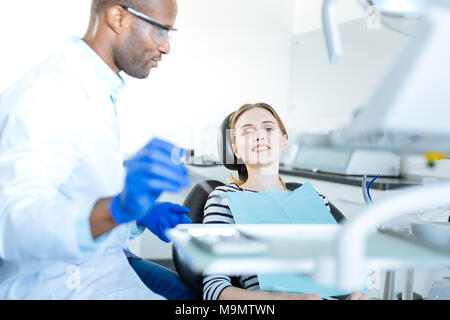 Bella paziente. Il focus è su un grazioso allegro giovane donna seduta in una poltrona del dentista e sorridente con la videocamera mentre si è in attesa di una cavità orale c Foto Stock