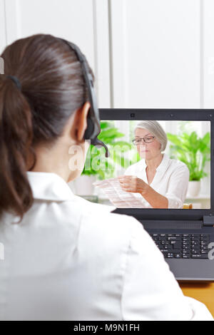 Il farmacista con cuffia nella parte anteriore del suo computer portatile durante una videochiamata con un paziente anziano tenendo il foglio di informazioni dei suoi farmaci su prescrizione Foto Stock