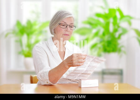 Senior donna seduta al tavolo nel suo soggiorno leggendo il foglio di informazioni della sua medicina prescritta Foto Stock