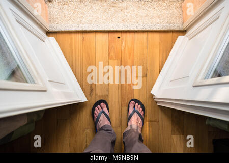 Buona mattina vacanze: Piedi di uomo di mezza età in flip-flop in piedi nella porta del balcone di un soleggiato rifugio per le vacanze Foto Stock