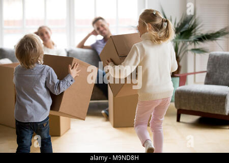 Vista posteriore a giocare i ragazzi con scatole sul giorno del trasloco, piccolo fratello e sorella in esecuzione tenendo averi imballaggio disimballato nella nuova casa, felice giocoso c Foto Stock