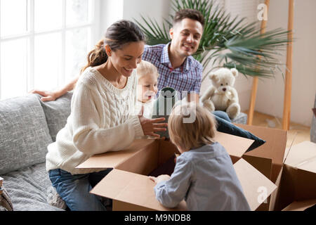 Felice famiglia con due bambini di disimballare le scatole dopo trasferimento in movimento o di sedimentazione in un nuovo concetto di home, eccitati i bambini piccoli per aiutare i genitori con b Foto Stock