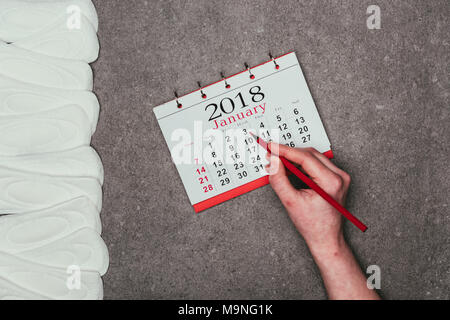 Ritagliato colpo di donna puntando alla data nel calendario con pastiglie mestruale intorno sulla superficie grigia Foto Stock