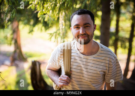 Felice l'uomo barbuto viaggiatore con zaino a piedi nella foresta. Turismo, Viaggi Avventura, escursione concetto - sorridente giovane uomo a camminare con zaino in legno Foto Stock