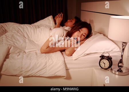 Giovane donna sbadigli nel letto mentre il marito tramite telefono cellulare accanto a lei di notte Foto Stock