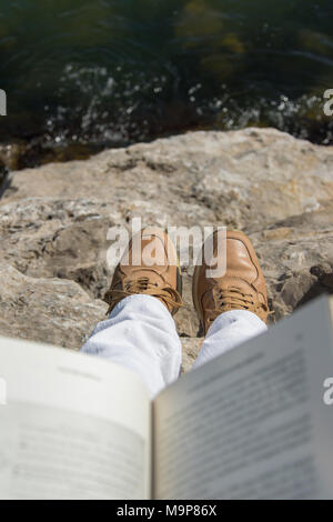 Vista al di sopra della mano di un adulto azienda libro aperto, seduta sulle rocce del mare Foto Stock