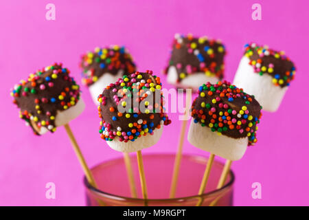 Marshmallows su una stick su sfondo rosa. vetrate con cioccolato e granelli colorati. Foto Stock