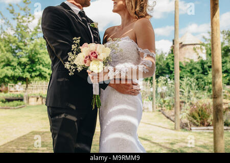 Sposa e lo sposo in piedi insieme con un mazzo di fiori il giorno delle nozze. Ritagliato colpo di sposa giovane in piedi all'esterno. Foto Stock