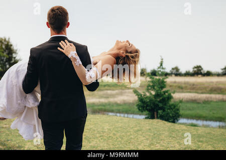 Colpo di dietro dello sposo che trasportano sposa nelle sue braccia a piedi. Vista posteriore della sposa giovane all'aperto nel parco. Foto Stock