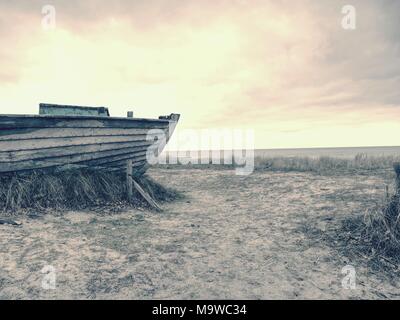 Abbandonate la barca disastrate bloccati nella sabbia. Vecchia barca in legno sulla riva sabbiosa della spiaggia. Tramonto sulla spiaggia. Vegetazione rada. Foto Stock