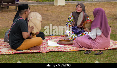 Un uomo Malay indossando un sonkok e tre donne che indossano hijabs sono seduti su un tradizionale tappeto di rattan e riproduzione del kompang (tamburo Malay). Foto Stock