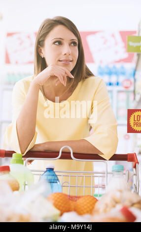 Attraente giovane donna fare la spesa al supermercato, ella è appoggiata sul carrello e di pensiero Foto Stock