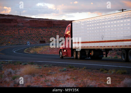 Big Rig americano moderno rosso semi carrello flotta con frigorifero semi rimorchio andando su strada tortuosa con rocce rosse colline del paesaggio dell'Arizona in twiligh Foto Stock