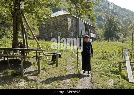 Una donna anziana cammina su un percorso sterrato dalla sua casa in pietra su una collina boschiva nelle montagne del Caucaso, Svaneti superiore, Georgia Foto Stock