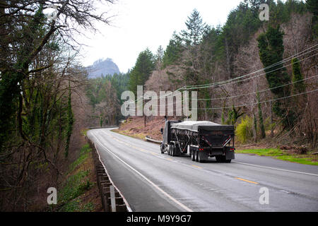 Classica cabina giorno americano nero big rig semi carrello trasporto coperto da tarp carichi alla rinfusa con il bulk semi rimorchio spostando sulla tortuosa strada in inverno Foto Stock