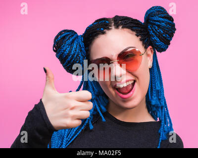 Entusiastico motivati attraente giovane donna con trecce blu acconciatura dando un pollice in alto gesto di approvazione e di successo con un sorriso raggiante. La messa a fuoco a portata di mano. Studio shot. Foto Stock