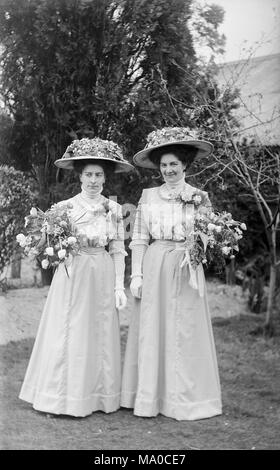 Due donne abbigliate come damigelle in fine l'Inghilterra vittoriana. Entrambi indossare abiti bianchi con elaborati bianco cappelli fiorito, ed entrambi portano una grande posy di fiori. Foto Stock