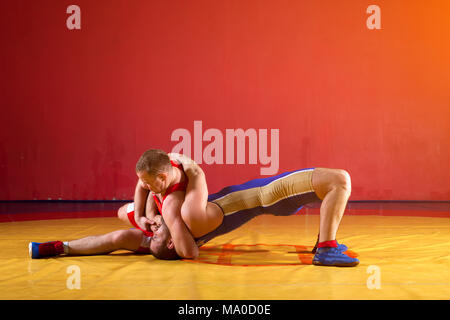 Due greco-romane lottatori in rosso e blu di wrestling uniforme su un giallo tappeto di wrestling in palestra Foto Stock