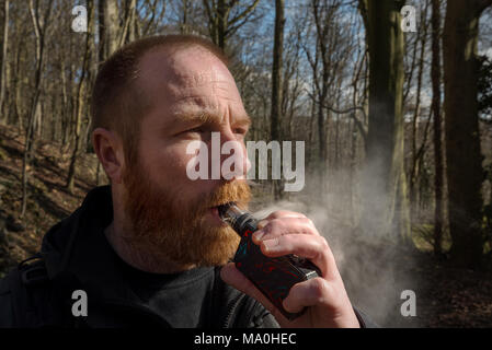 Lo zenzero un uomo barbuto vapes su una casella mod con vetro vaporizzatore. Foto Stock