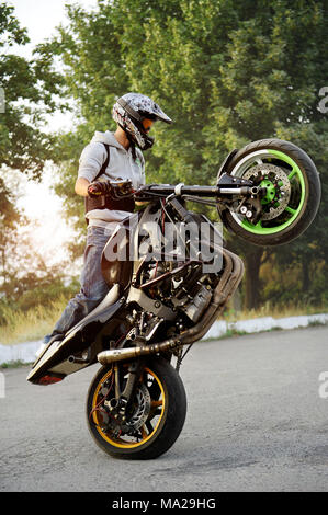 Ivano-Frankivsk, Ucraina - 28 Agosto 2015 : buona foto di uno stuntman facendo trick riding motociclo su un ciclo lungo la strada. Trucchi verde sullo sfondo. Tramonto d'estate. Foto Stock