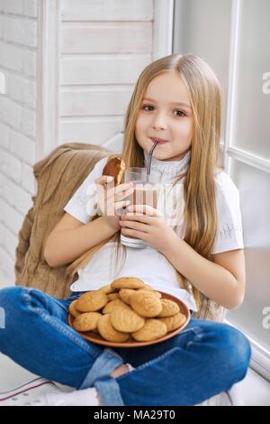 Bambina è seduta su una sedia,jeans blu e bianco felpa. Bambino beve latte al cioccolato da vetro trasparente ed ha grande lastra piene di dolci deliziosi biscotti sulle sue ginocchia. Foto Stock