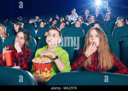 Tre amici mangiare popcorn da guardare film. Ragazza di bere bevanda frizzante dal big red può. I bambini a guardare i nuovi film o cartoon. cercando interessati e abbandonata. Indossare magliette colorate. Foto Stock