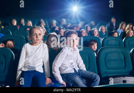 Frontview del ragazzo e ragazza seduta nella sala cinema e guardando i nuovi film o cartone animato. Ragazzo sta guardando la telecamera, ragazza è la visione di film su schermo cinematografico. abbandonata, emotiva bambini sullo sfondo. Foto Stock