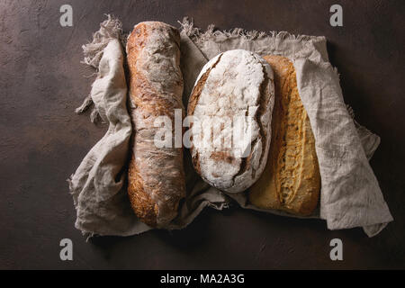 Varietà di polpettine sfornato fresco artigianale di segale, bianco e grano intero pane sulla tela di lino su marrone scuro dello sfondo texture. Vista superiore, copia dello spazio. Foto Stock