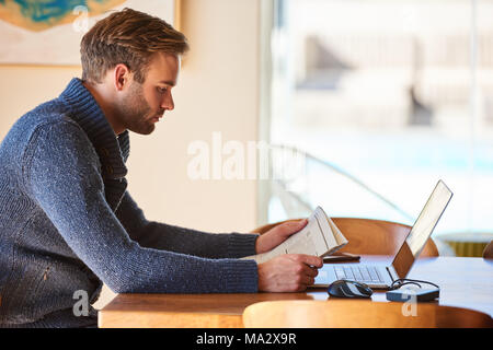 Bianco uomo seduto al tavolo del soggiorno al mattino presto occupato a leggere il suo quotidiano per recuperare sugli affari correnti, con la sua tazza di caffè Foto Stock