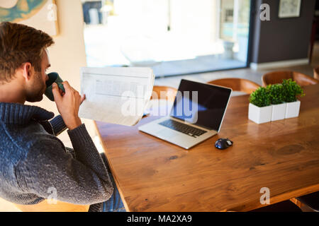 Uomo caucasico prendendo un sorso di mattina tazza di caffè durante la lettura del quotidiano a suo bel tavolo da pranzo, con il suo notebook davanti a lui per w Foto Stock