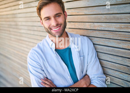 Strettamente immagine ritagliata del bel giovane maschio nella sua metà 20s sorridente in telecamera con i suoi denti e mostrando le braccia incrociate mentre appoggiata contro beauti Foto Stock