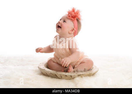 Un sorridente, sette mesi di età bambina indossa una pesca colorata, romper a maglia. Lei è seduta in una ciotola di legno. Girato in studio su un tappeto sheepkin. Foto Stock