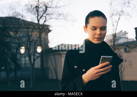 Giovane donna utilizza smart phone mentre permanente sulla strada. Lei indossa mantello nero e sorrisi mentre texting. Foto Stock