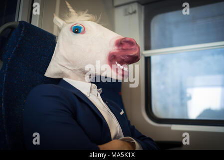 Insolito passeggero in elegante abito si siede da solo all'interno del treno e pronto a viaggiare. Giovane uomo gravi in maschera comica. Funny unicorn traveler Foto Stock