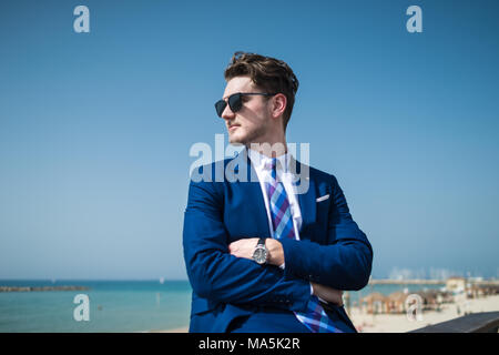 Giovane uomo gravi in elegante abito e sunglass sullo sfondo del mare e del cielo. Manager di successo all'aperto rilassante nel giorno caldo e soleggiato. Ragazzo alla moda Foto Stock