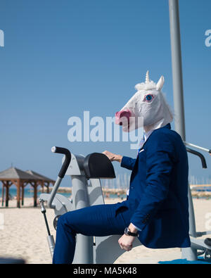 Strano ragazzo in tuta e maschera non esercizi a sport terra vicino alla spiaggia. Uomo insolito muscoli delle pompe. Funny unicorn è impegnato nel fitness all'aperto Foto Stock