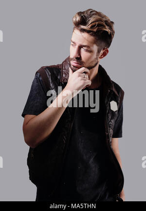 Perplesso giovane uomo accarezzare un toccante barba, guardando la telecamera su uno sfondo grigio Foto Stock