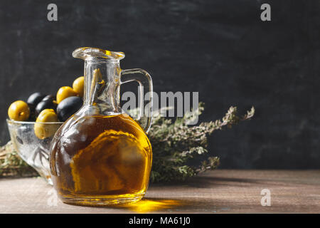 Bottiglia di vetro del premio di olio vergine di oliva, ciotola con olive ed erbe su sfondo scuro, stile rustico Foto Stock