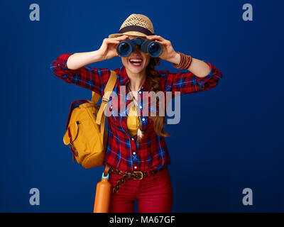 La ricerca di luoghi ispiratrice. Sorridendo turistica attiva donna con zaino guardando attraverso il binocolo isolato su blu Foto Stock