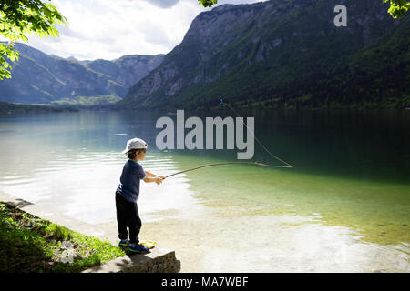 Giovane ragazzo in piedi sulla riva e pesca con uno in legno realizzati a mano polo di pesca sul lago di Bohinj, sulle Alpi Giulie, Slovenia Foto Stock