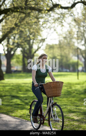 Giovane donna attraente cavalca la sua bicicletta attraverso un soleggiato parco a Londra. Foto Stock