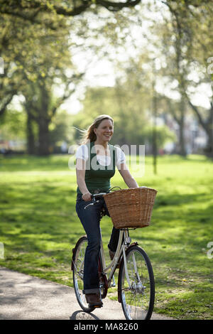 Giovane donna attraente cavalca la sua bicicletta attraverso un soleggiato parco a Londra. Foto Stock