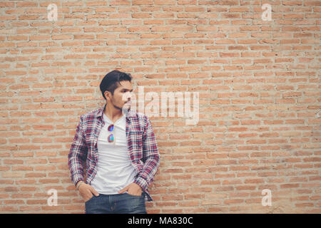 Giovane bello hipster uomo che indossa plaid shirt e jeans contro un muro di mattoni. Foto Stock