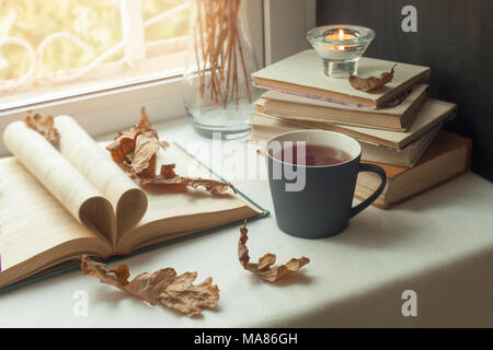 Autunno still life. libri, foglie, cuo di tè e asciugare minuscoli fiori in vaso rosso candelabro con una candela che brucia sulla finestra. Concetto di lettura di autunno Foto Stock