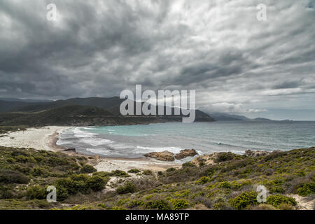 Costa rocciosa del deserto des Agriates e Ostriconi spiaggia in Corsica con Ile Rousse nella distanza sotto il grigio cielo tempestoso e macchia mediterranea in foregr Foto Stock