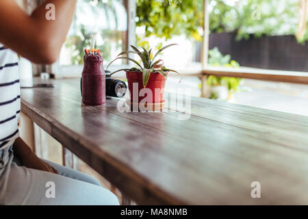 Donna seduta accanto a una finestra con un smoothie sul tavolo. Vaso Smoothie collocato su un tavolo accanto a una fotocamera e un vaso di fiori. Foto Stock