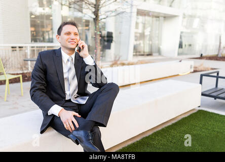 Bello e attraente giovane impreditore seduta sul banco di lavoro, usando parlare tenendo lo smartphone mobile telefono cellulare sorridente in tuta e cravatta, allegro su Foto Stock