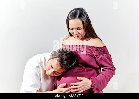 Attraente giovane donna incinta indossando un bordeaux abito senza maniche seduto mentre guardando verso il basso in corrispondenza di suo marito che è in ascolto di un bambino con la sua e Foto Stock